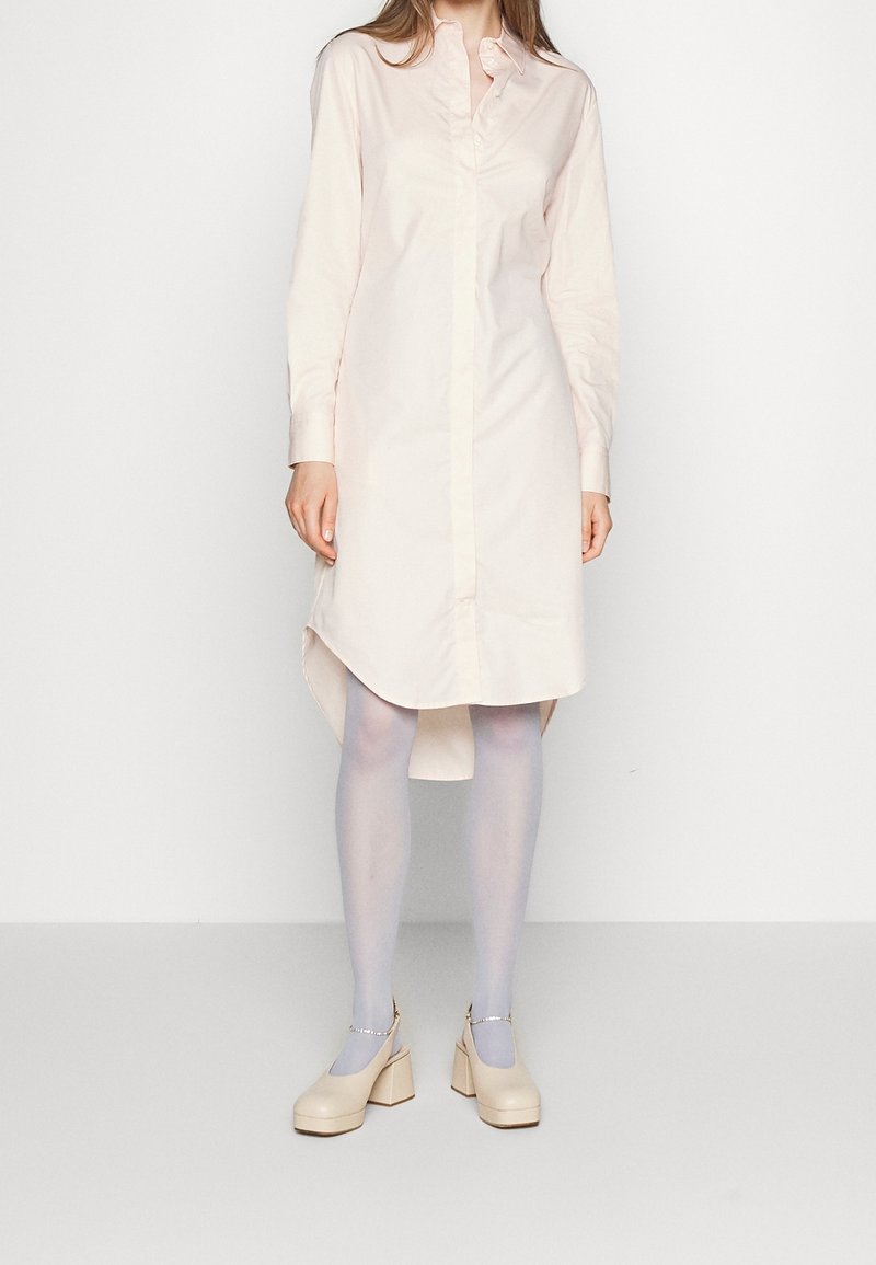 Mujer que lleva un vestido camisa de manga larga color rosa pálido, medias azul claro y zapatos Mary Jane beige con tacón de bloque y correas en el tobillo.