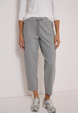 Personne portant un pantalon de survêtement court gris clair avec des poches zippées et des baskets argentées, debout contre un mur beige uni.
