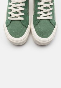 Zapatillas de lona verdes con suelas y cordones de goma blanca. Presentan un upper texturizado, costuras en contraste y un diseño de puntera redonda.