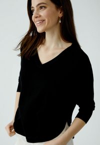 Schwarzer V-Ausschnitt-Pullover mit drei Viertelärmel. Weiche Textur, seitliche Schlitze und lockere Passform. Über einer hellen Unterbekleidung getragen.