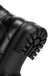 Czarna skórzana boot z grubą, teksturowaną gumową podeszwą, charakteryzującą się głębokimi wzorami bieżnika zapewniającymi przyczepność oraz gładką górą z bocznymi detalami szwów.