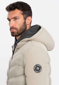 Giacca puffer beige con cappuccio, dotata di una fodera interna grigia, texture trapuntata e un patch circolare con logo sulla manica sinistra.