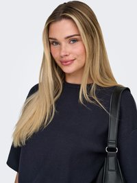 Mlajša ženska z dolgimi svetlimi lasmi, oblečena v temno mornarsko srajco, rahlo se smehlja in nosi črno usnjeno torbico na desnem rami.