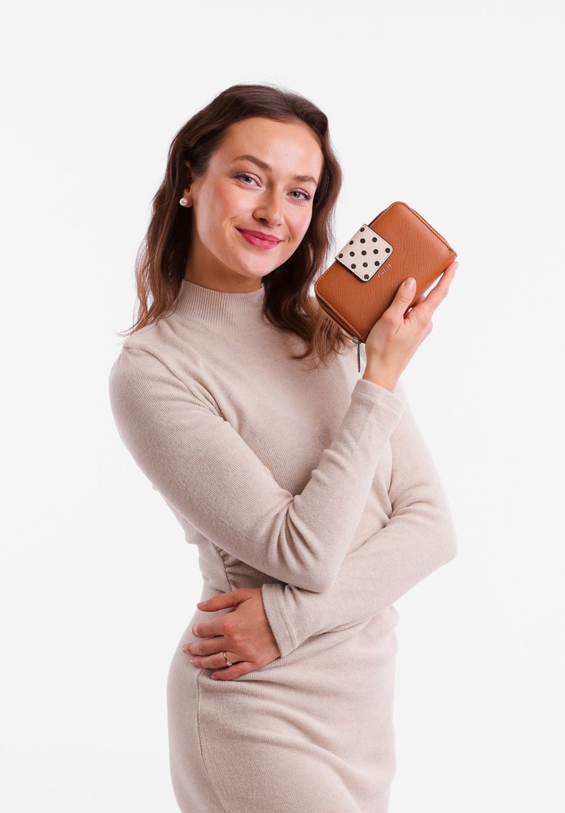 Brown leather wallet with a white polka dot accent and a zip closure, held by a person wearing a light beige sweater in a plain background.