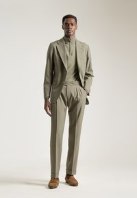 Traje verde oliva con chaqueta a medida y pantalones con pliegues, combinado con una blusa ajustada de alto cuello y mocasines de ante marrón.