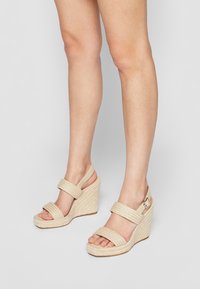 Beige kile-sandaler med rebtekstur; har to brede stropper over foden og en slingback-strop med en metalspænde-accent.