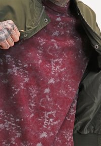 Capa de suéter de forro polar con estampado rojo y gris debajo de una chaqueta bomber verde oliva, con cierre delantero y puños acanalados.
