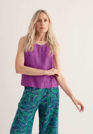 Vrouw met lang blond haar, gekleed in een mouwloze paarse top en groene broek met bladmotief tegen een effen achtergrond.