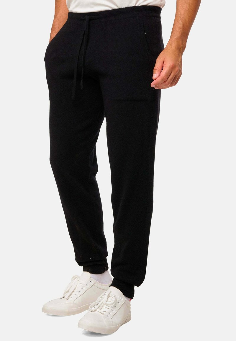 Homme portant un pantalon de jogging noir avec des chevilles élastiques et un cordon de serrage, associé à des baskets blanches et des chaussettes blanches.