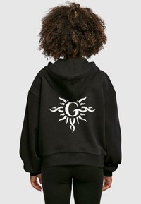 Fekete, oversized kapucnis pulóver, amelyen egy nagy fehér napminta található a hátsó részen egy középen lévő "G" betűvel. Az anyag lágy hatású, laza illeszkedéssel.