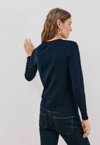 Granatowy sweter z długim rękawem, okrągłym dekoltem i guzikami na rękawach, połączony z ciemnoniebieskimi dżinsami. Gładka tekstura.