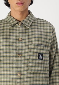 Grön och beige rutig skjorta med knäppt krage, träknappar och en bröstficka med en svart märkeslapp.