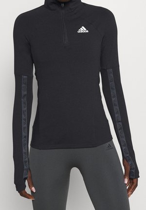 Haut Adidas noir à manches longues avec demi-fermeture éclair, logo sur la poitrine et rayures à motif sur les manches, porté avec des leggings gris Adidas.