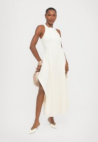 Witte mouwloze jurk met een hoge halslijn en zijsplits, gemaakt van textiel met een structuur. Gecombineerd met crèmekleurige schoenen en een beige tas.
