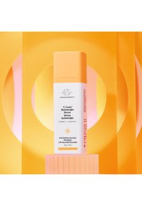 White serum bottle with orange cap, featuring text about hydration. Set against bright yellow and pink geometric backgrounds.