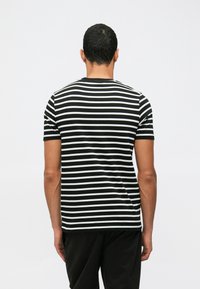 Vyras stovi atsukęs nugarą kamerai, vilkėdamas juodai baltą horizontalių raštų trumpomis rankovėmis marškinėlį ir juodas kelnes.