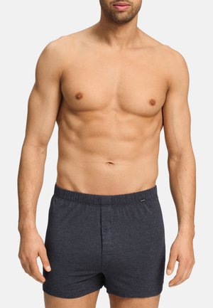 Hombre sin camiseta, con pantalones bóxer de cintura elástica gris oscuro, de pie con los brazos relajados frente a un fondo claro liso.