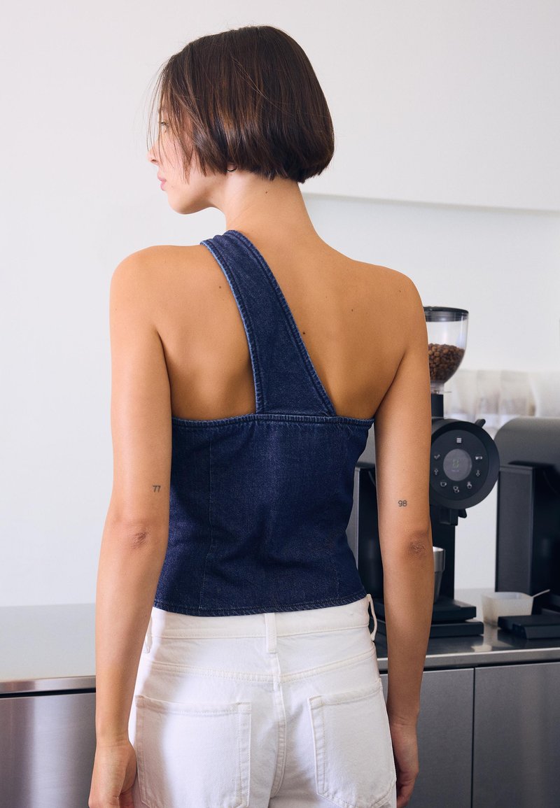 Kvinde med kort hår, set bagfra, iført en mørkeblå, enkelt skulder top og hvide bukser, stående ved siden af en kaffekværn i et køkken.