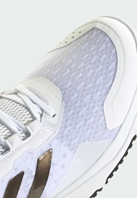 Białe sportowe buty z teksturowaną cholewką, z metalicznymi złotymi akcentami, sznurówką i trwałą czarną podeszwą.