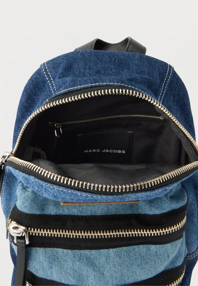 Σακίδιο πλάτης Denim Marc Jacobs με ανοιχτό φερμουάρ στην κορυφή, που αποκαλύπτει το μαύρο εσωτερικό και εσωτερική τσέπη με φερμουάρ και ετικέτα της μάρκας.