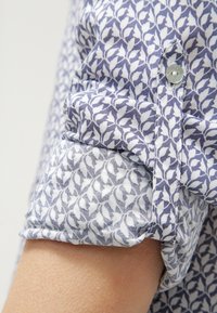 Camicia con colletto e maniche arrotolate, caratterizzata da un motivo a foglia blu e bianco su morbido tessuto di cotone. Dettaglio dei bottoni al polsino.