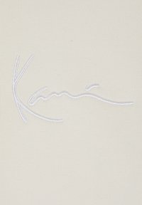 Krémová tkanina s výrazným bielym vyšívaným podpisom v plynulej písaní. Textúra je hladká s jemným žíhaním.