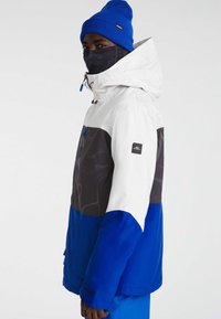 Kleurjack met lagen voor de winter; heeft witte, zwarte en levendige blauwe panelen. Soepele stof met een logo op de mouw en een capuchon.