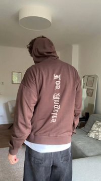 Smeđi dukserica s velikim bijelim vertikalnim tekstualnim motivom na leđima. Ima kapuljaču s vezicama i opušteni kroj. Uparena s sivim hlačama.