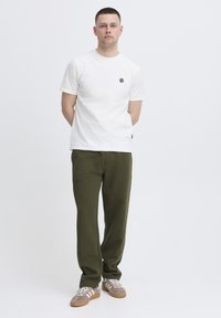 Hvid bomulds-T-shirt med en lille sort emble, kombineret med olivengrønne joggingbukser og grå sneakers med hvide striber. Simpelt design.