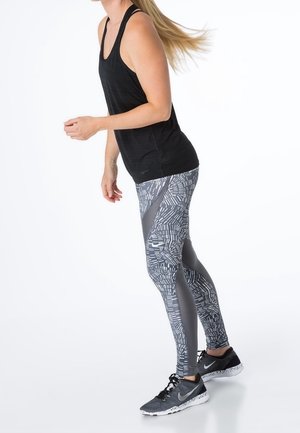 Top atlético negro combinado con leggings grises estampados que presentan diseños geométricos. La modelo lleva zapatillas negras con detalles blancos.