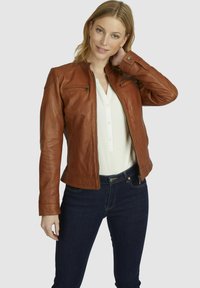 Chaqueta de cuero marrón con un diseño ajustado, bolsillos con cremallera y un cuello alto. Combinada con una blusa de color claro y unos vaqueros oscuros.