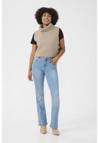 Bézs bordás kötött ujjatlan garbó crop top, három barna gombbal az oldalon, világoskék, egyenes szárú farmerral és bézs cipővel párosítva.