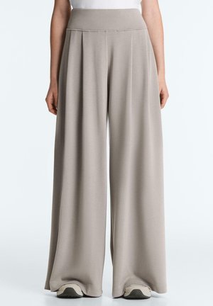 Kobieta w beżowych szerokich spodniach z wysokim stanem i plisami, zestawionych z białymi trampkami i białą bluzką na jednolitym tle.