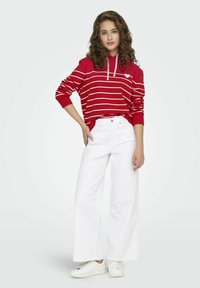 Felpa a righe rosse con strisce bianche e una tasca frontale, abbinata a jeans bianchi a gamba larga e sneakers bianche. Capelli ricci e posa rilassata.