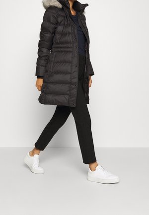 Abrigo puffer negro con capucha forrada de piel, con textura acanalada, botones a presión y bolsillos laterales. Combinado con pantalones negros y zapatillas blancas.