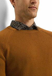 Brauner, strukturierter Pullover mit einem gerippten Muster, kombiniert mit einem schwarzen, blumenmusterbesetzten Hemd mit Kragen, das weiße und orange Blumen zeigt.