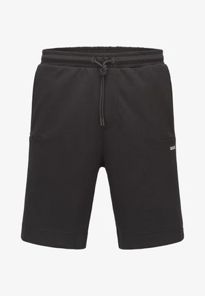 Hombre con sudadera negra con cremallera, camiseta blanca, pantalones cortos negros, calcetines blancos y zapatillas blancas, de pie contra un fondo gris liso.
