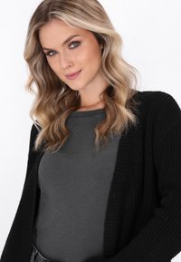 Donna bionda con capelli ondulati, che indossa un top grigio scuro a costine e un cardigan nero, sorridendo dolcemente su uno sfondo neutro.