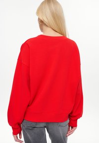Persoon met lang blond haar die een helderrood oversized sweatshirt en grijze jeans draagt, staand met de rug naar de camera tegen een effen witte achtergrond.