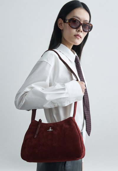 Bolsa de camurça vermelha com uma textura suave, uma forma estruturada, uma alça curta e um acabamento em metal prateado. Modelo vestindo uma camisa branca e uma gravata.