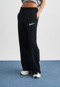 Črne trenirke z ohlapnim krojem, z zategovalnim trakom v pasu in srebrnim odsevnim logotipom Nike na levi stegni. Mehak teksturni material.