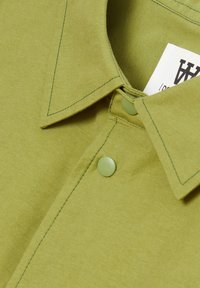 Green collared shirt with a smooth texture, featuring contrasting stitching and a light green button at the neck. Care label visible.