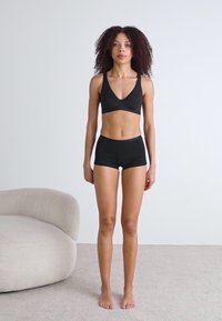 Ensemble de haut de sport noir et de shorts, présentant un design simple avec une texture lisse. Le modèle se tient sur un tapis clair près d'un canapé courbé.