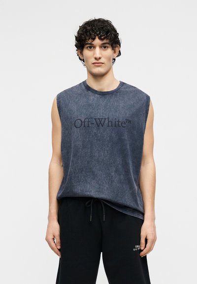 Νέος με σγουρά μαλλιά, φοράει αμάνικο ξεθωριασμένο μαύρο t-shirt Off-White και μαύρο παντελόνι με κορδόνι, σε απλό φόντο.