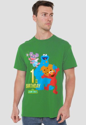 Grünes Baumwoll-T-Shirt mit Cartoonfiguren: Elmo, Krümelmonster und Abby Cadabby. "1. Geburtstag"-Text in Gelb. Rundhalskragen.