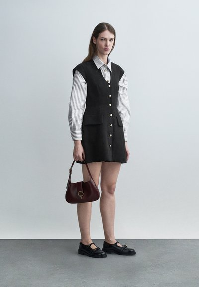 Femme en robe noire boutonnée sans manches par-dessus une chemise à rayures, tenant un sac à main rouge foncé, portant des chaussures Mary Jane noires, debout sur un sol gris.