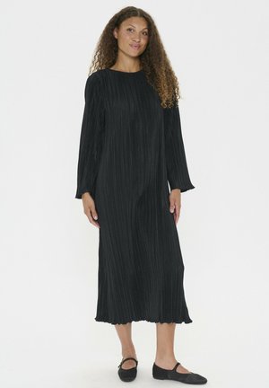 Langes schwarzes, gefälteltes Kleid mit langen Ärmeln und rundem Halsausschnitt; der Saum hat eine gewellte Kante. Das Model trägt schwarze flache Schuhe.