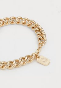 Zelta ķēdes aproce ar bieziem, savijamiem posmiem un mazu gravētu tagu, kas pievienots aizdarei. Gluda, spīdīga virsma ar atstarojuma efektu.