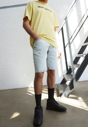 Gul bomuld t-shirt med teksten "MUSIC TELEVISION", lyseblå denimshorts, sorte crew-sokker og sorte sneakers.