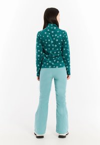 Mujer de pie de espaldas, vistiendo una chaqueta de forro polar de color teal con patrón de estrellas, pantalones de nieve azul claro y botas blancas, sobre un fondo blanco.
